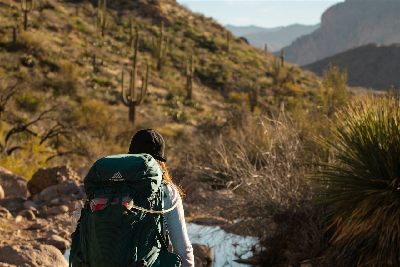 A prepared hiker with a backpack walks a clear desert trail past saguaro cacti in the cool morning light
