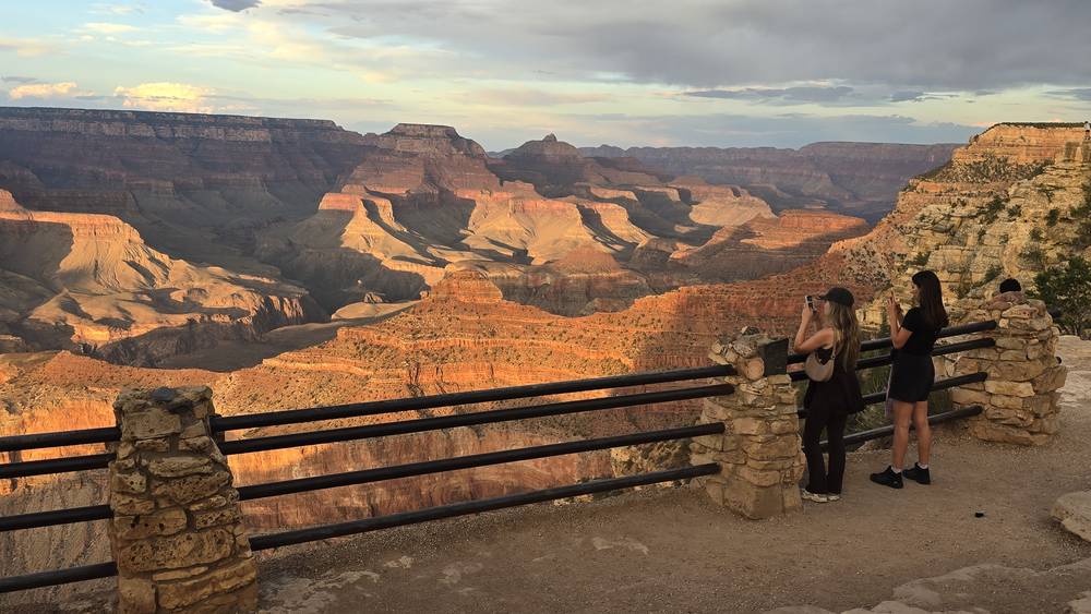 Two visitors photograph the Grand Canyon from a stone-walled South Rim overlook as the canyon glows in late afternoon light